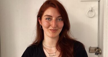 Basically a long headshot of a smiling woman standing against a white wall.