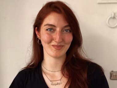 Basically a long headshot of a smiling woman standing against a white wall.