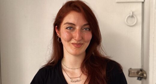 Basically a long headshot of a smiling woman standing against a white wall.
