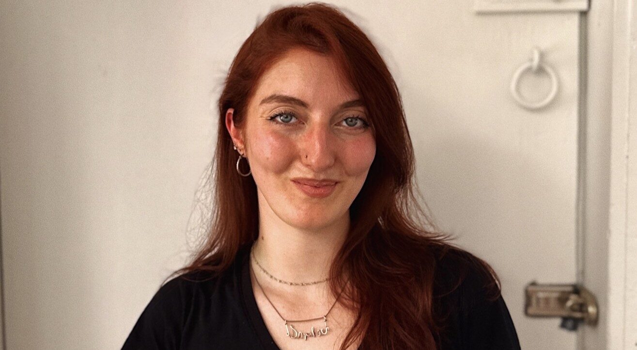 Basically a long headshot of a smiling woman standing against a white wall.