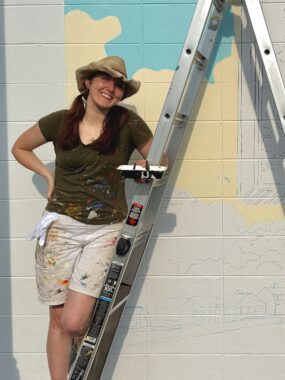 A woman in a cowboy hat smiles on a hot, sunny day (she's sweating) whil standing against a freshly painted wall and behind a step ladder. 