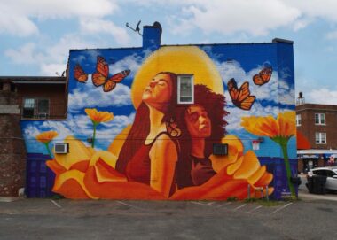 A mural that takes up the side of a building shows bright orange, red, blue, and white colors depicting two women standing back to back, eyes closed, and looking up to a blue sky with white, puffy clouds and large monarch butterflies. 