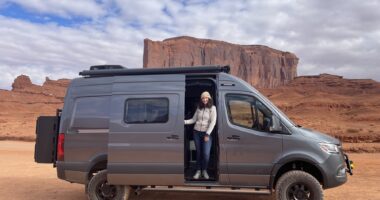 A woman stands inside the doorway of a van seen against a backdrop of large rock formations.