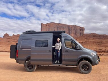 A woman stands inside the doorway of a van seen against a backdrop of large rock formations.