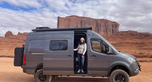 A woman stands inside the doorway of a van seen against a backdrop of large rock formations.