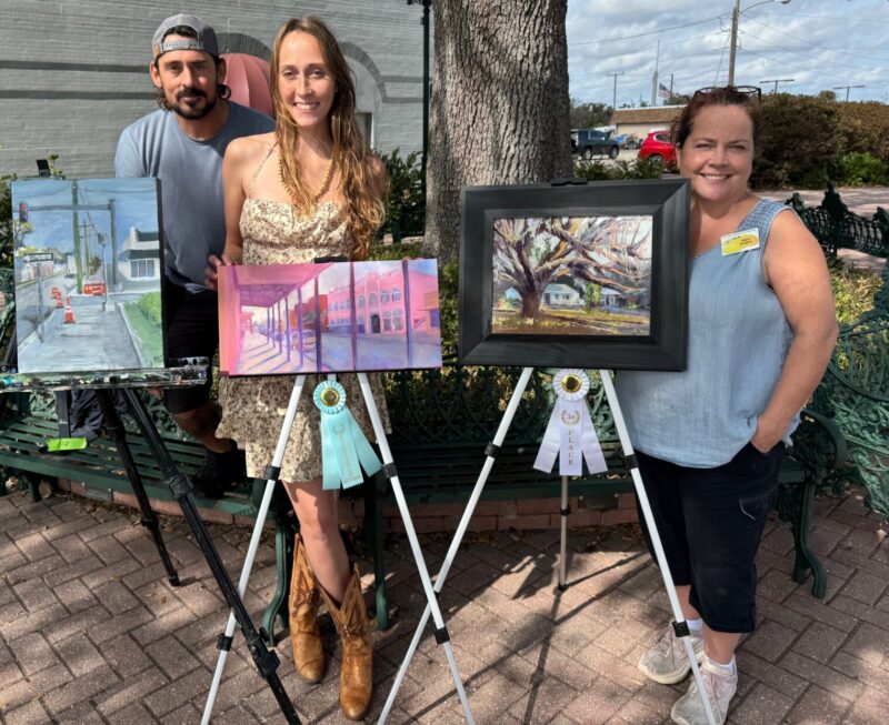 A man and two women stand behind three portable art easels, each with small paintings on them. The two women's paintings have prize ribbons attached to them. 