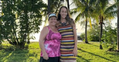 Two women - mother and daughter - stand and pose on a vast lawn in front of the beach under palm trees on a sunny day.