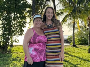 Two women - mother and daughter - stand and pose on a vast lawn in front of the beach under palm trees on a sunny day.