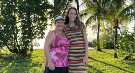 Two women - mother and daughter - stand and pose on a vast lawn in front of the beach under palm trees on a sunny day.
