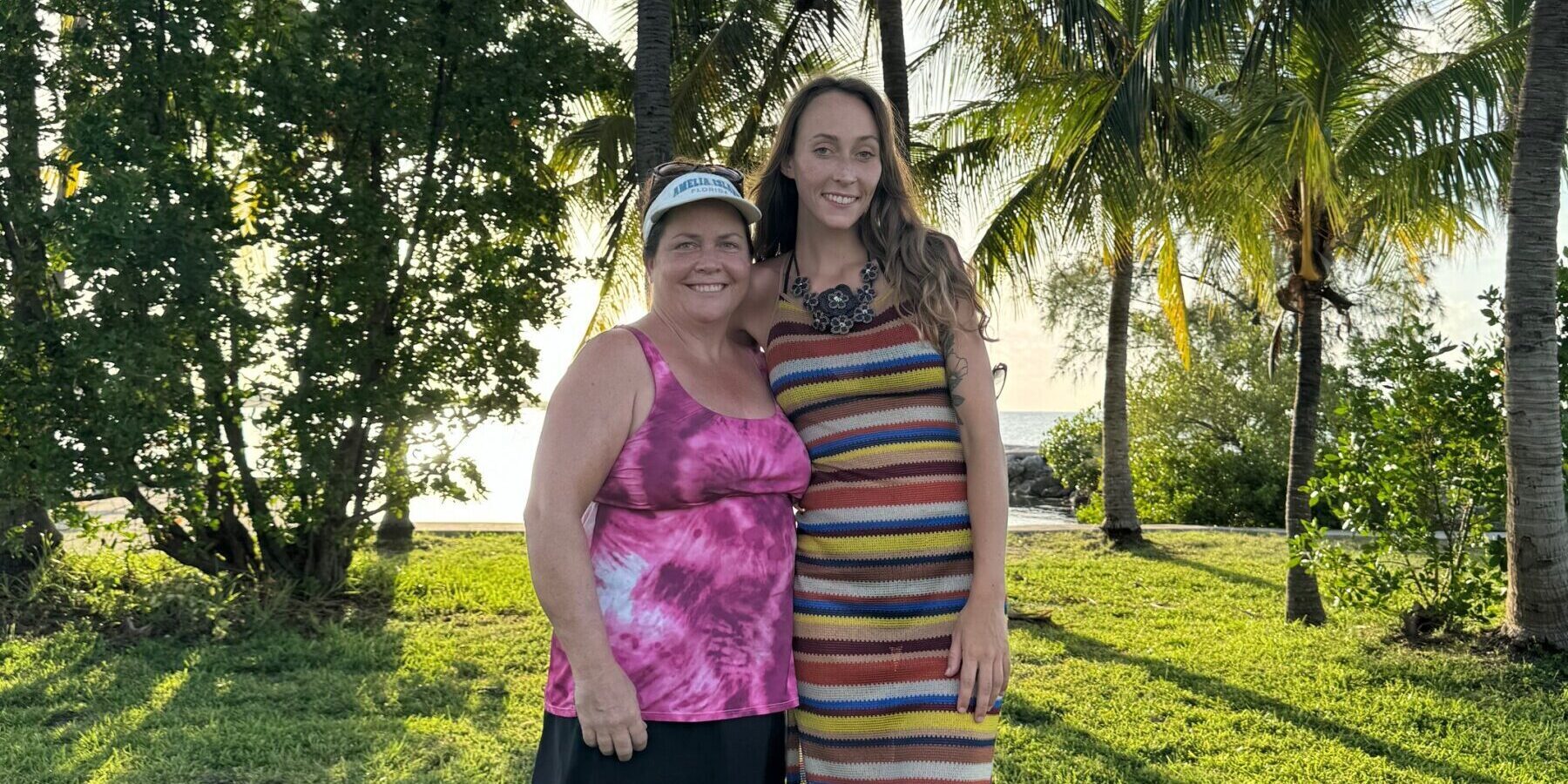 Two women - mother and daughter - stand and pose on a vast lawn in front of the beach under palm trees on a sunny day.
