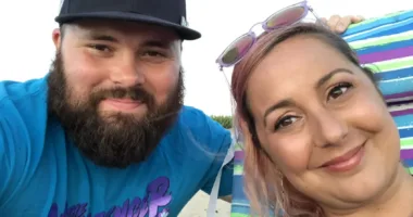 A close-up of a man and a woman smiling. The bearded man is wearing a Boston Red Sox cap and the woman has sunglasses resting on her head.