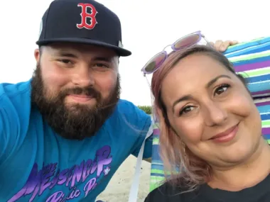 A close-up of a man and a woman smiling. The bearded man is wearing a Boston Red Sox cap and the woman has sunglasses resting on her head.