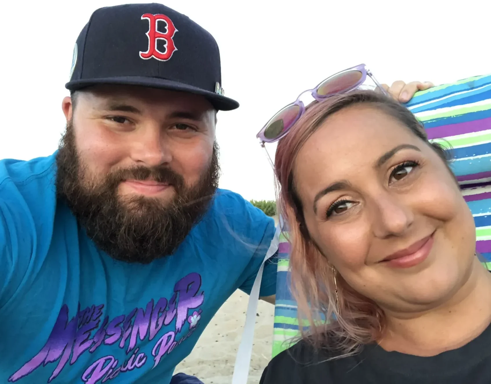 A close-up of a man and a woman smiling. The bearded man is wearing a Boston Red Sox cap and the woman has sunglasses resting on her head.