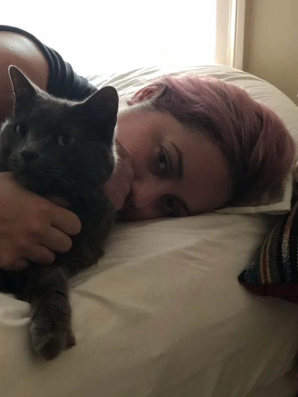 A close-up of a woman's head lying on a pillow in bed with her arm around a cat. 