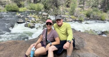 A photo shows a woman and man sitting on a rock in nature.