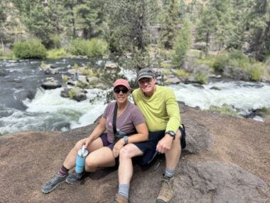 A photo shows a woman and man sitting on a rock in nature.