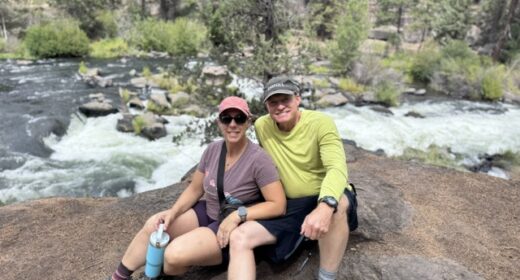 A photo shows a woman and man sitting on a rock in nature.