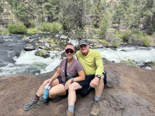 A photo shows a woman and man sitting on a rock in nature.
