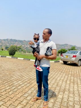 A man stands outside on what appears to be a driveway, holding his young daughter in his arms. She has on dark sunglasses and is reaching one hand out toward the camera.