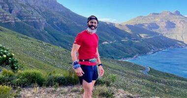 A man in a backward baseball cap and red shirt and blue shorts stands about midway up a gorgeous mountain that descends to a large, blue body of water. It's sunny and you can tell he's been exercising.