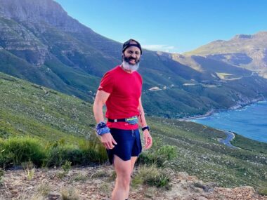 A man in a backward baseball cap and red shirt and blue shorts stands about midway up a gorgeous mountain that descends to a large, blue body of water. It's sunny and you can tell he's been exercising.