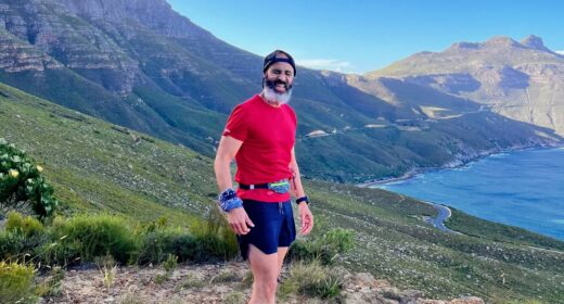 A man in a backward baseball cap and red shirt and blue shorts stands about midway up a gorgeous mountain that descends to a large, blue body of water. It's sunny and you can tell he's been exercising.