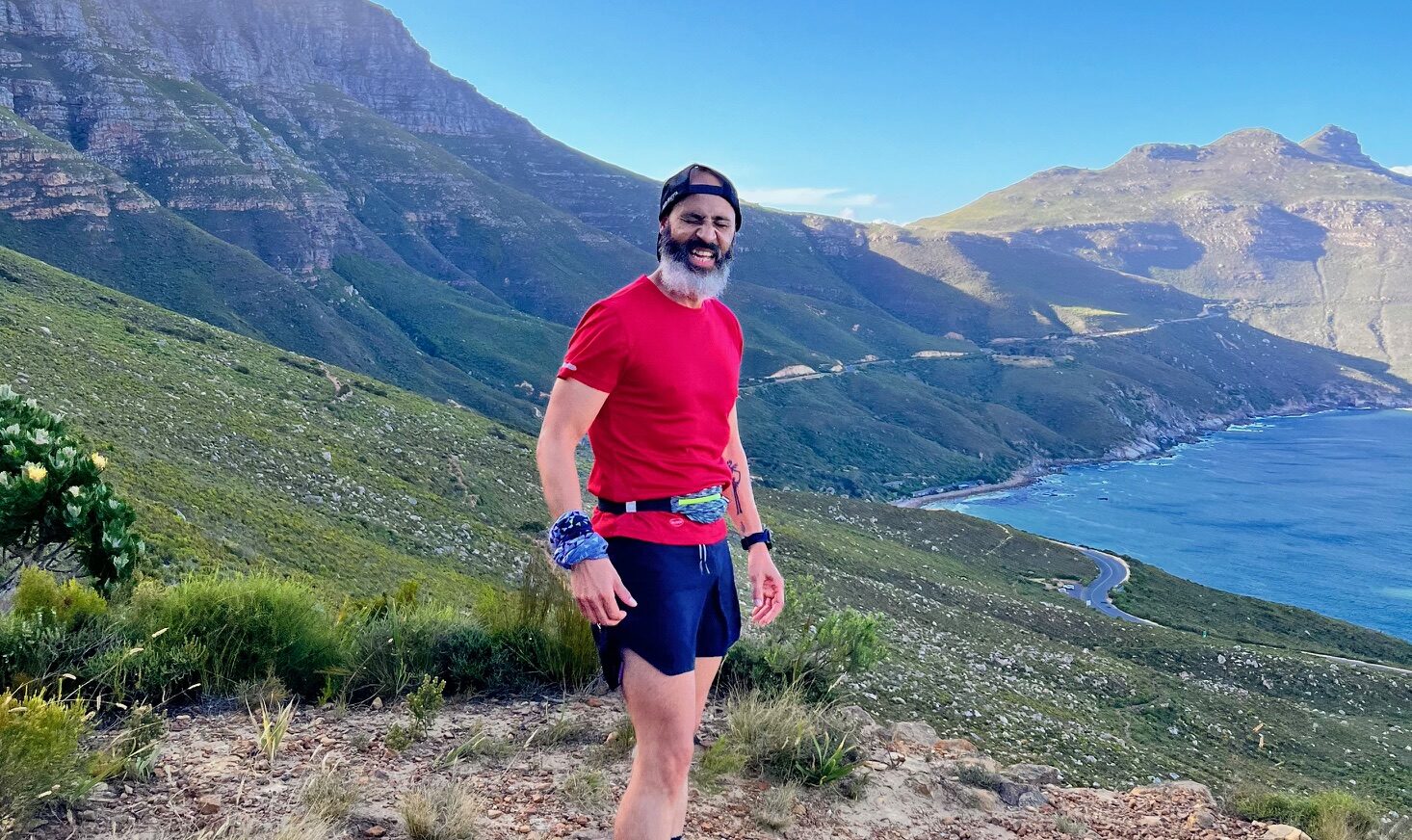 A man in a backward baseball cap and red shirt and blue shorts stands about midway up a gorgeous mountain that descends to a large, blue body of water. It's sunny and you can tell he's been exercising.