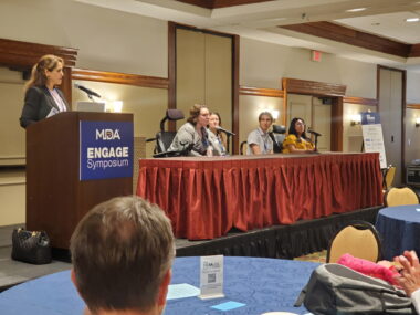 A woman stands at a podium with a plaque reading "MDA Engage." People are sitting behind a table to the right, where one person is speaking into a microphone.