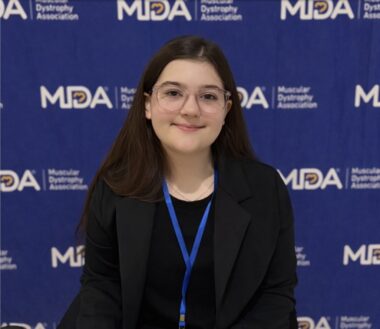 A 12-year-old girl wears a black blazer and a blue lanyard while standing in front of a backdrop featuring the MDA logo.