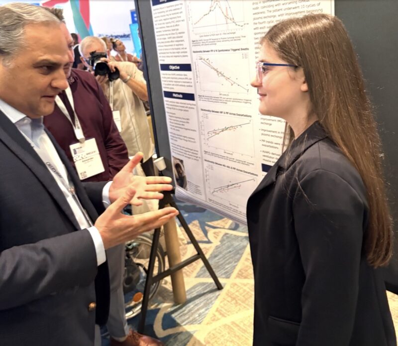 A 12-year-old girl wearing a black blazer is talking with an older man in front of a poster board featuring her research on respiratory monitoring in MG.